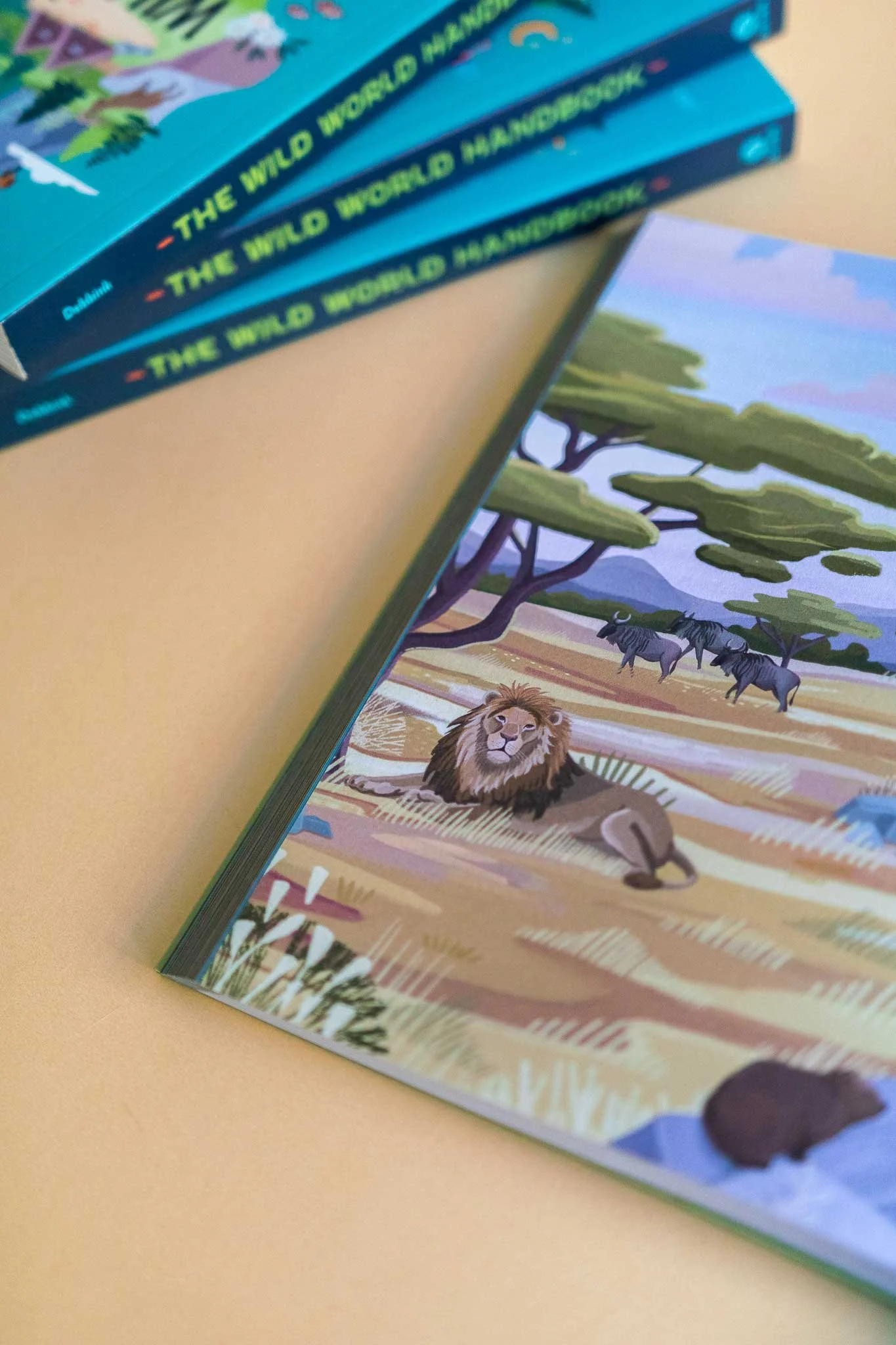 Open children's book with illustrated safari scene featuring a lion, zebras, and a hippopotamus, with additional books stacked beside it.