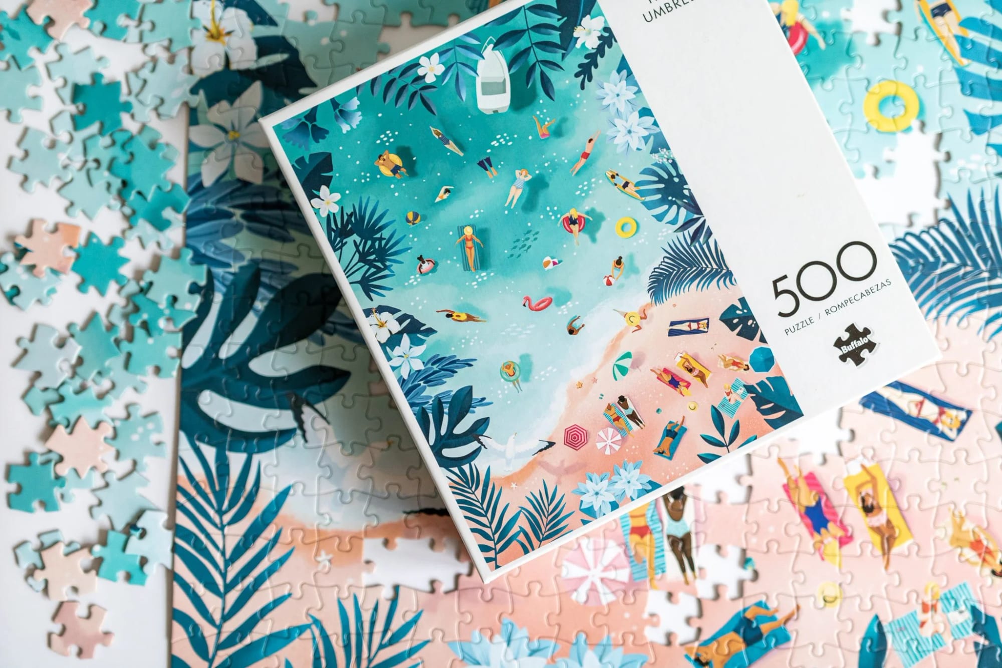 Puzzle box depicting a beach scene with people swimming, sunbathing, and relaxing under umbrellas, surrounded by tropical plants and flowers, with puzzle pieces scattered around.