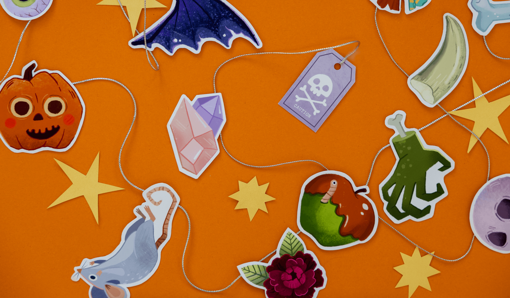 Halloween-themed paper cutouts and stickers on an orange background, including a pumpkin face, bats, stars, a ghost, a skull, a green slime hand, a rose, a worm in an apple, a bone, a gemstone, and caution tags.