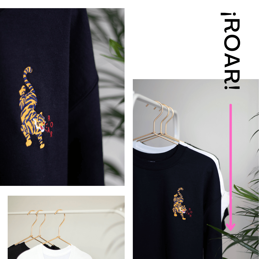 Close-up of a navy sweatshirt with an embroidered yellow and black tiger and the word 'ROAR' on a hanger, with other sweatshirts hanging nearby and green plants in the background.