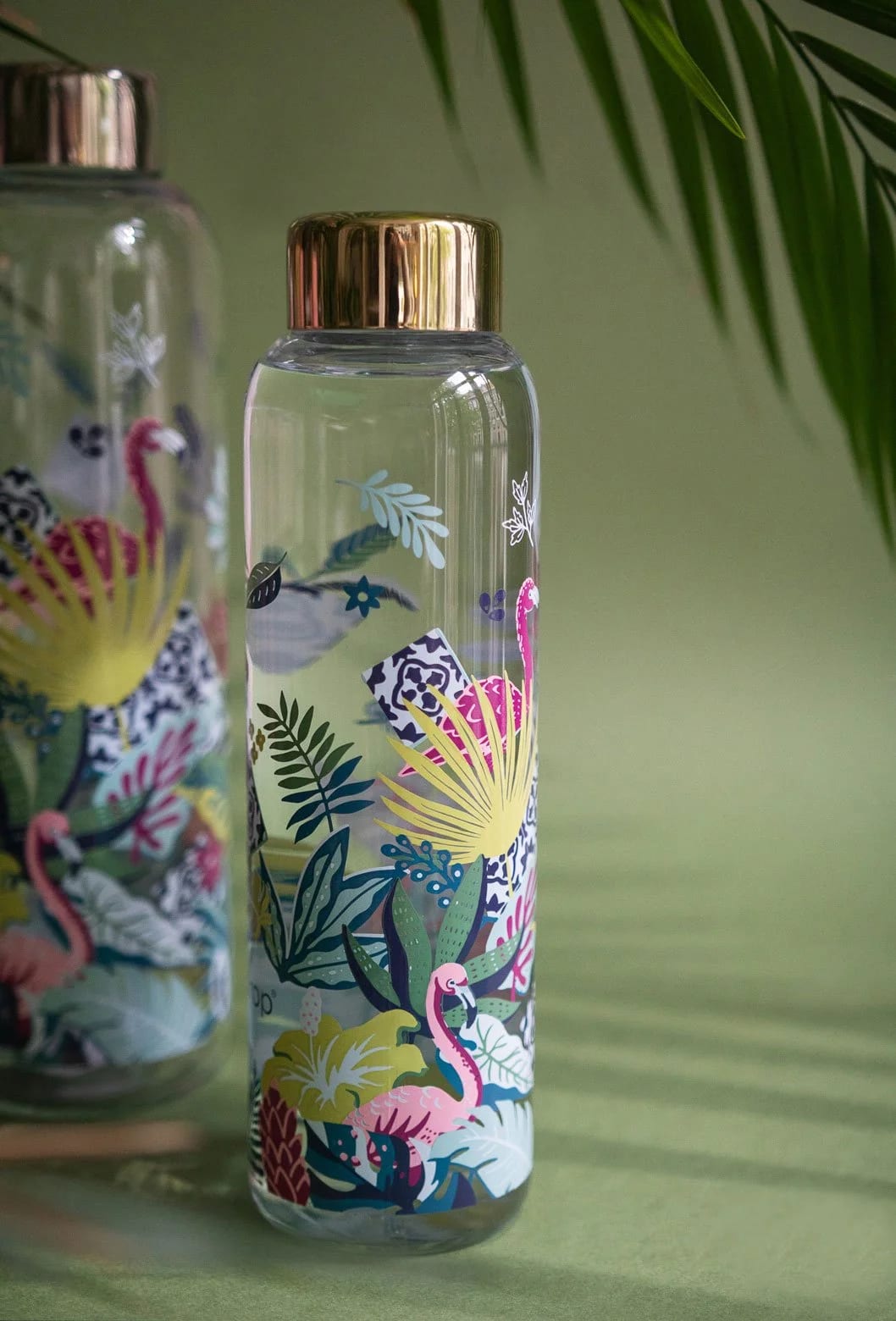A clear glass water bottle with a gold-colored lid, featuring colorful tropical flamingo and leaf patterns, placed on a green surface with green leafy plants in the background.