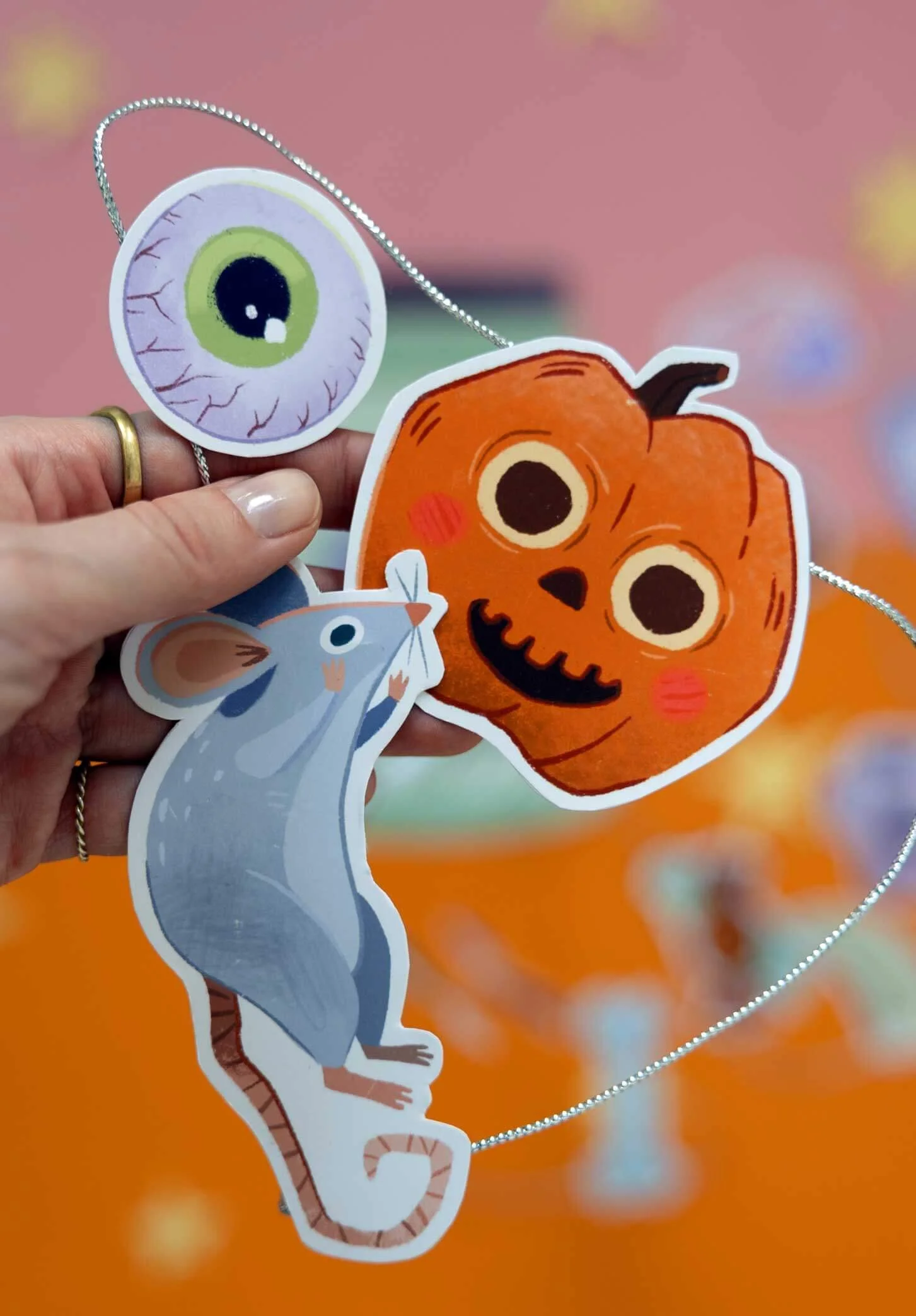 Hand holding three Halloween-themed paper cutouts of an eyeball, a pumpkin with a carved face, and a gray mouse, connected with a string, against an orange background with blurred decorations.