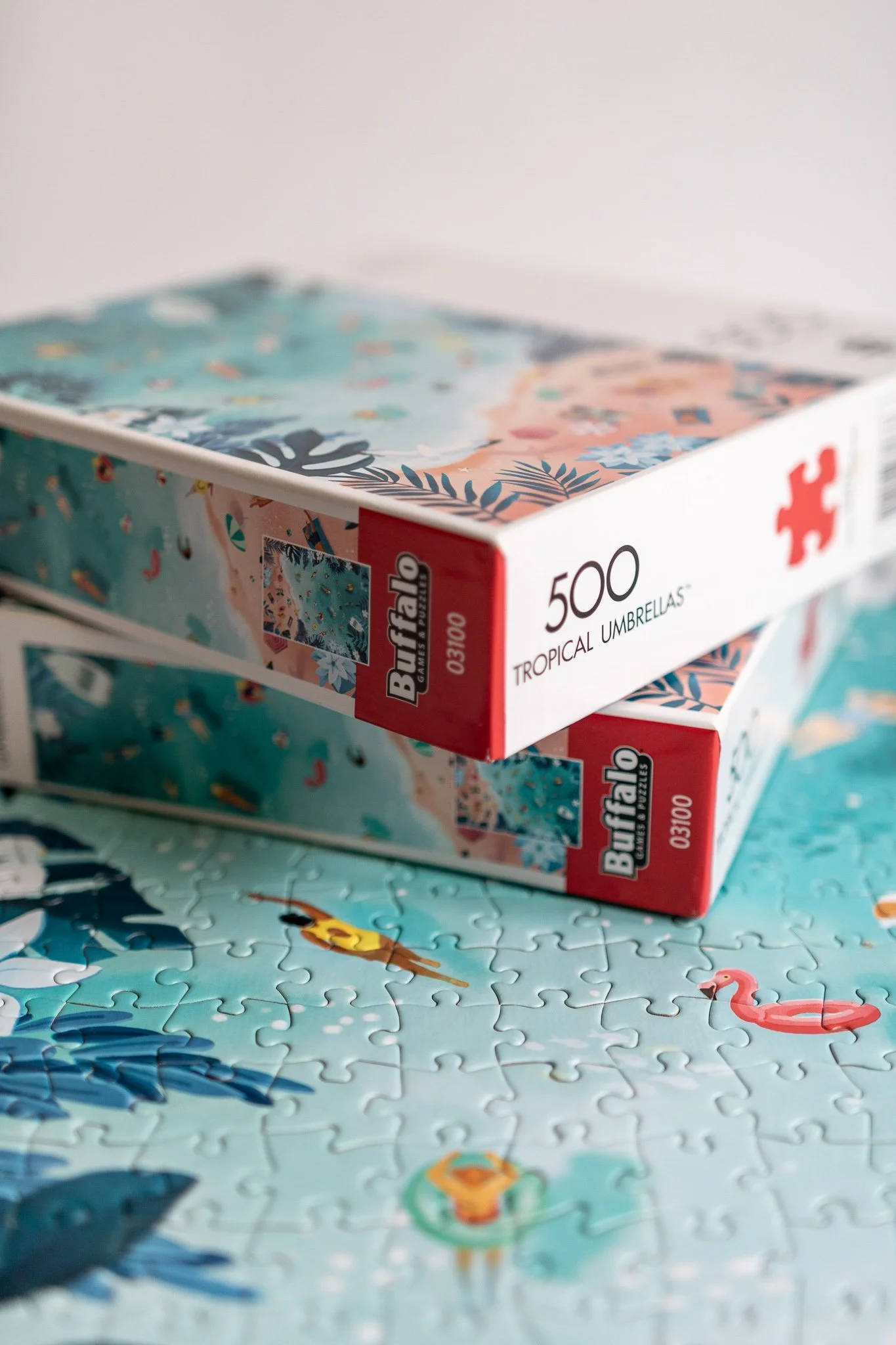 Two boxes of Buffalo 500 tropical umbrellas resting on a completed puzzle with a beach scene, including a flamingo float and palm trees.