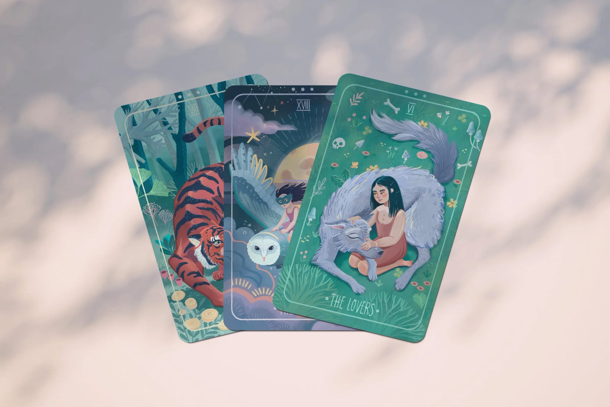 Three illustrated tarot cards with nature and animal themes. The card on the right, titled 'The Lovers,' depicts a woman with long black hair sitting beside a large gray wolf in a forest setting. The middle card shows a fairy and owl under a starry night sky. The left card features a tiger in a jungle environment.