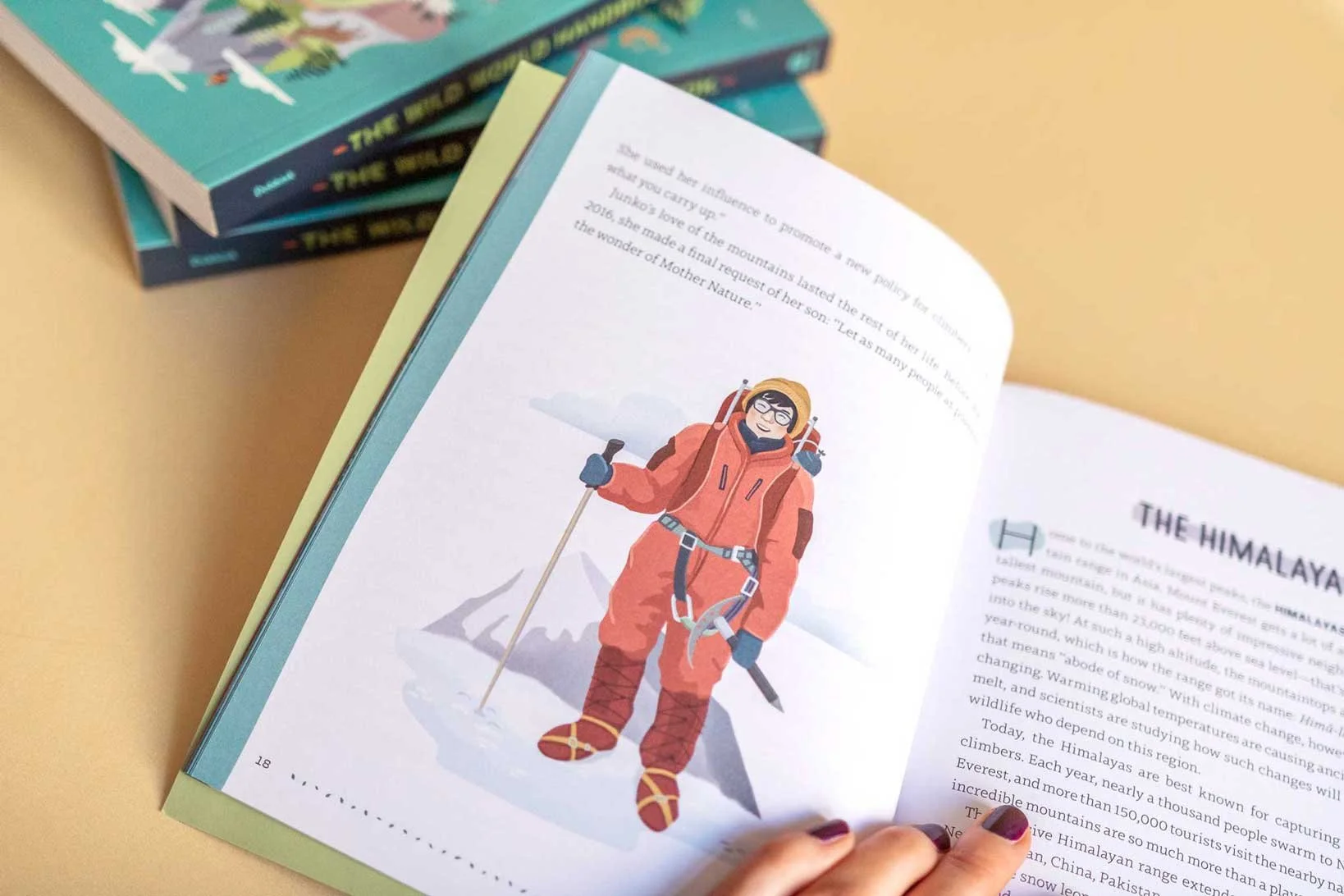 Open book showing an illustration of a person in orange mountaineering gear standing on snow with mountains in the background, holding a trekking pole, wearing gloves, glasses, and crampons.