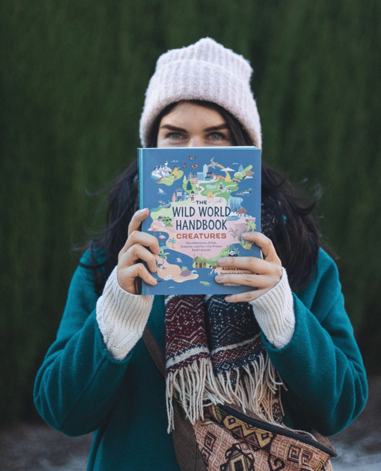 A woman wearing a pink knit hat and a teal coat holding a book titled 'The Wild World Handbook: Creatures' in front of her face, standing outdoors with trees in the background.