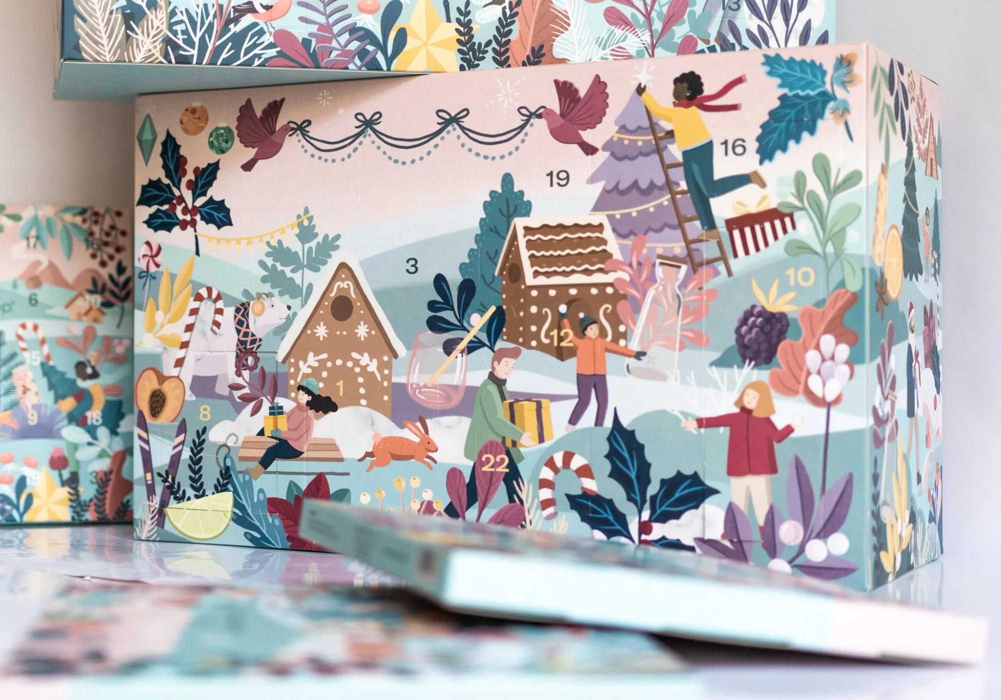 Illustrated Christmas scene on an advent calendar box featuring children decorating a tree, wrapping gifts, and playing in a snowy landscape with gingerbread houses, holly, and berries.