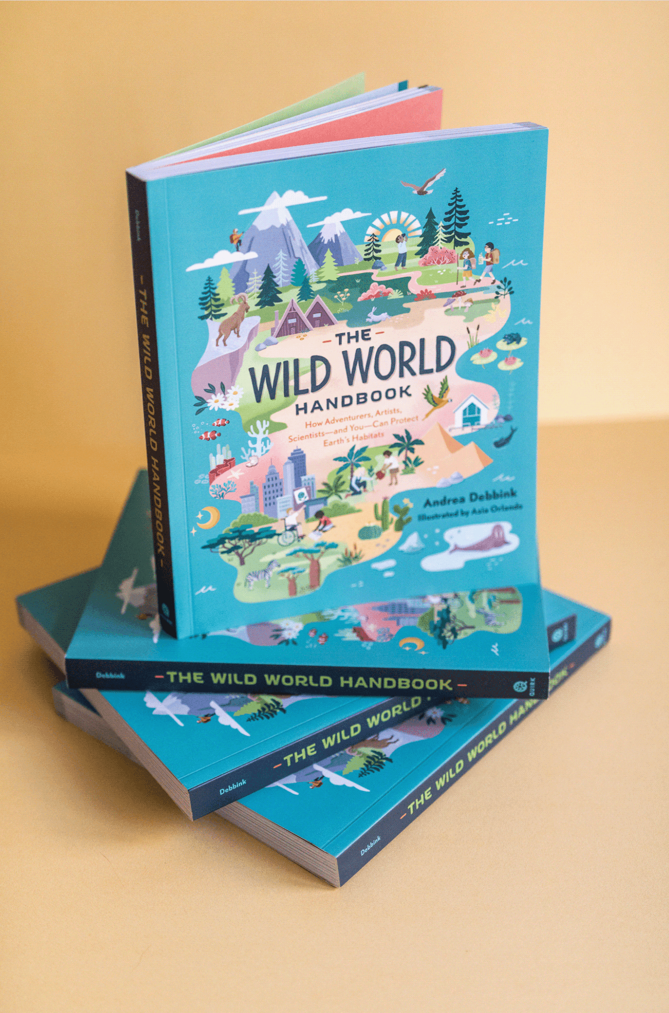 Three copies of 'The Wild World Handbook' with a colorful illustrated cover featuring nature scenes, mountains, trees, animals, and people, placed on a yellow surface.