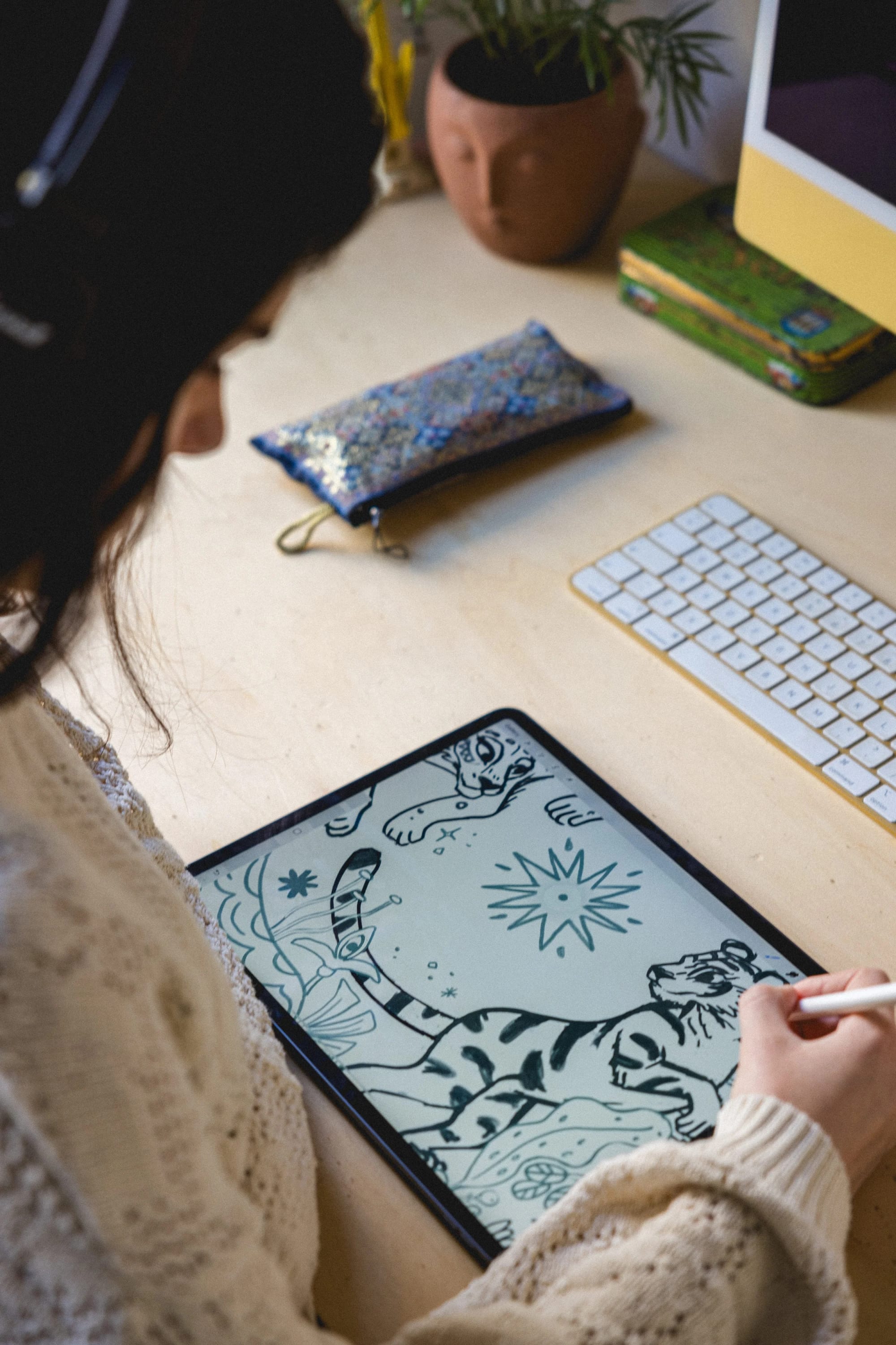 A person drawing a black and white sketch of a tiger on a tablet, sitting at a desk with a keyboard, a colorful clutch, and decorative items including a plant and a face-shaped pot.