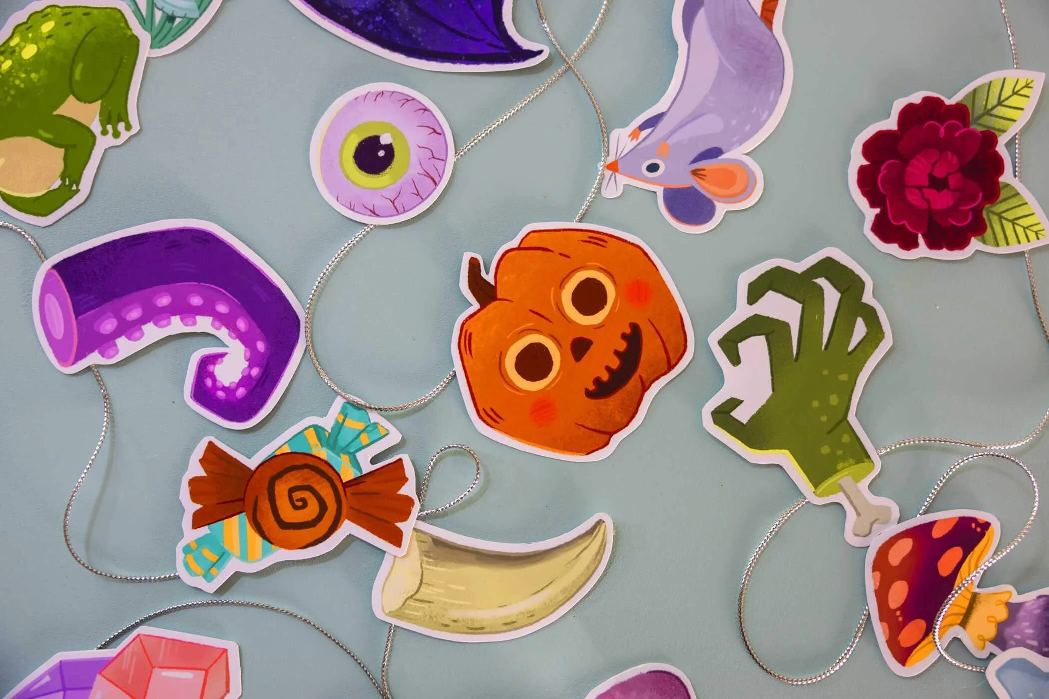Halloween-themed paper stickers and jewelry, including a pumpkin, purple tentacle, eyeball, frog, butterfly, flower, mushroom, hand, banana, candy, and other decorative elements on a light background.