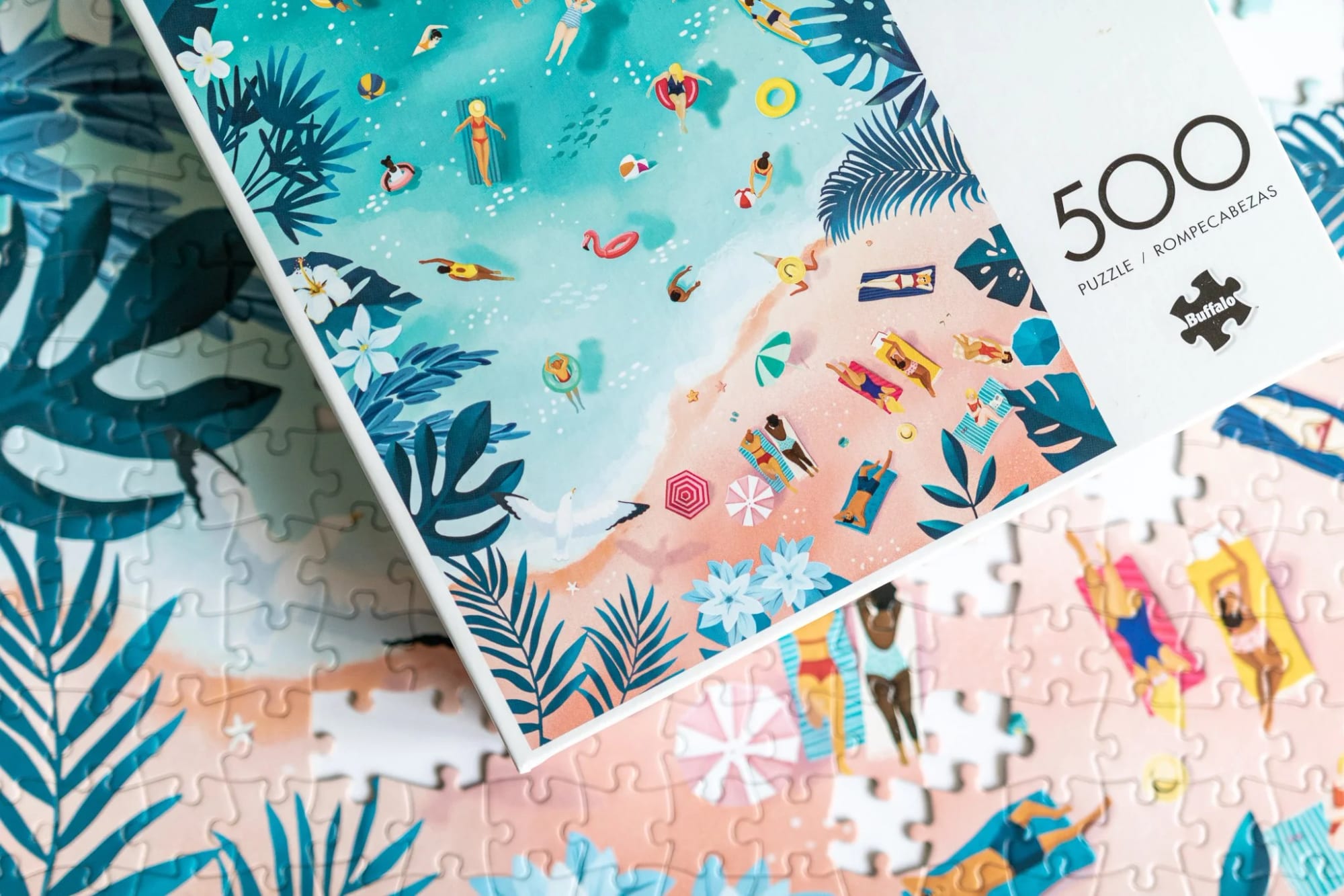 A puzzle box featuring a colorful illustration of a beach scene with people swimming, sunbathing, and relaxing on the sand, surrounded by tropical foliage and flowers.
